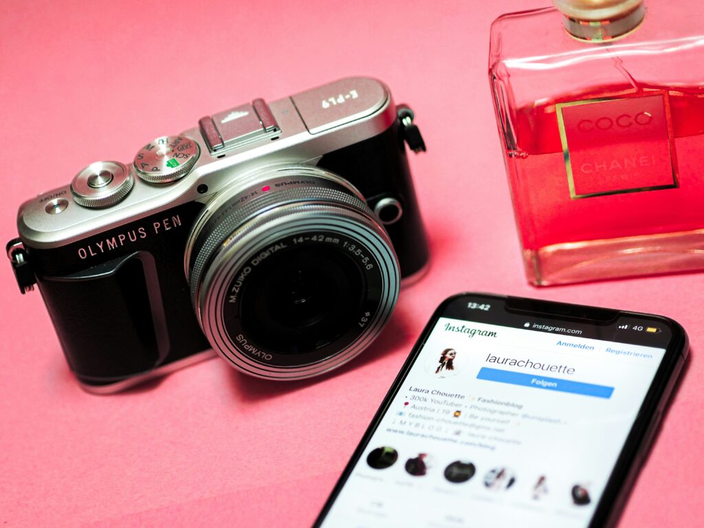 A camera and a perfume with a pink background placed next to a phone displaying an influencer's profile.