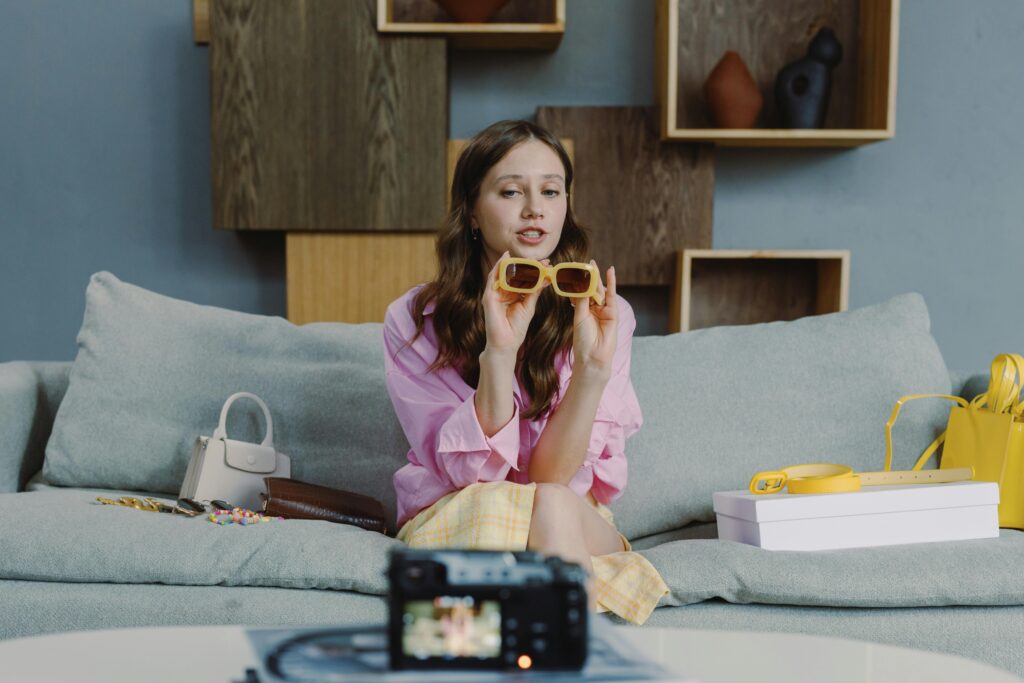 A woman is sitting on a sofa in front of a camera recording user-generated content for a sunglasses brand.