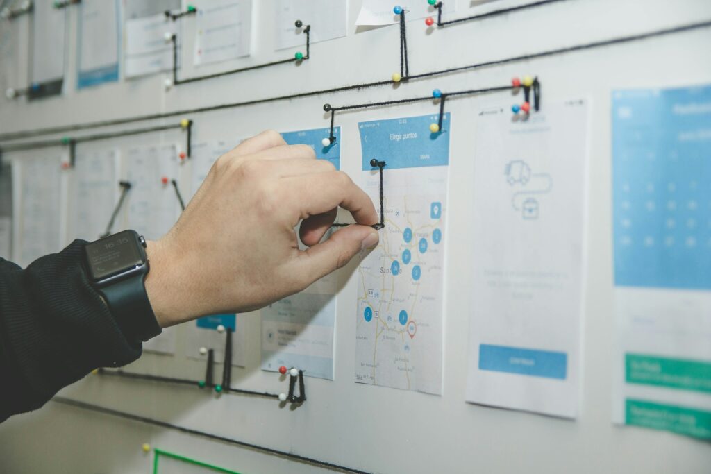 A hand wearing a smartwatch arranges pinned paper mockups on a wall, connected by a string diagram.