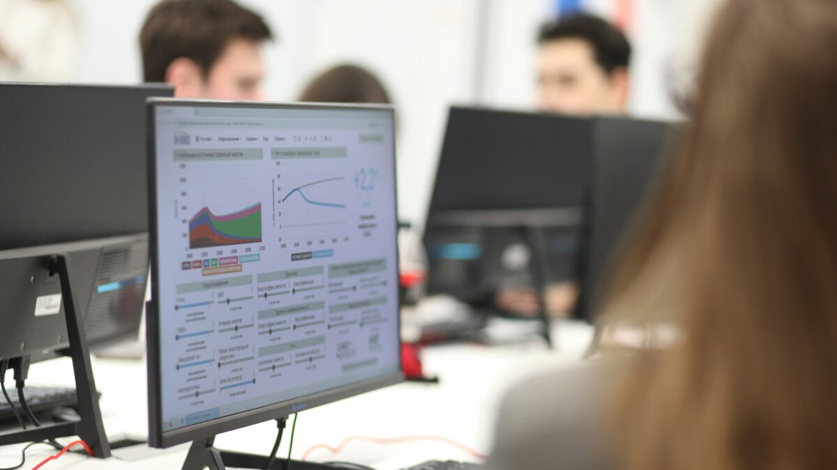 Office setting with people working at computers. A monitor displays colorful graphs and data charts.