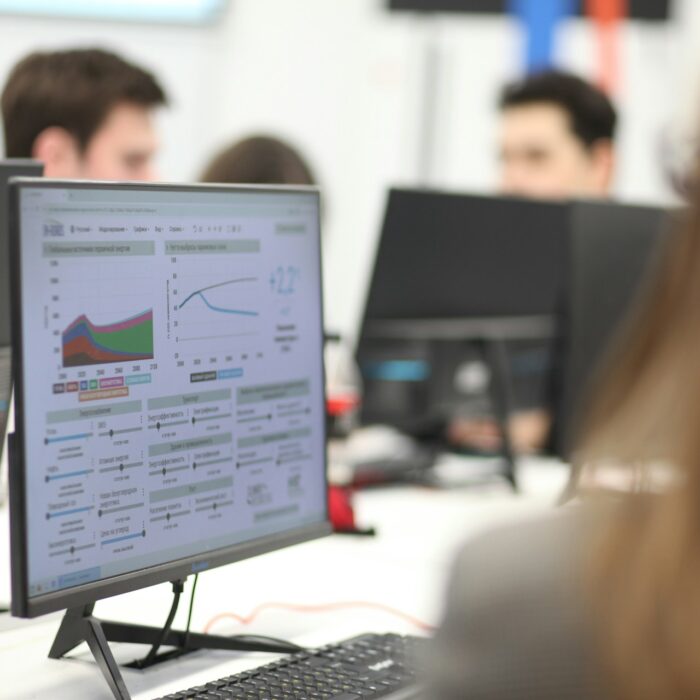 Office setting with people working at computers. A monitor displays colorful graphs and data charts.