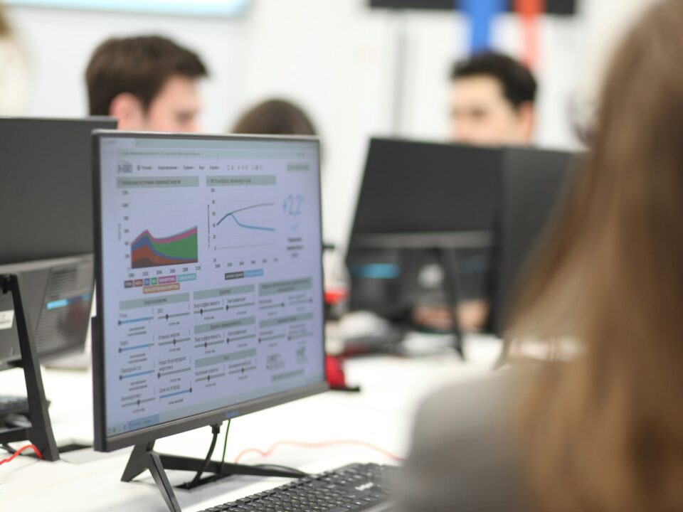 Office setting with people working at computers. A monitor displays colorful graphs and data charts.