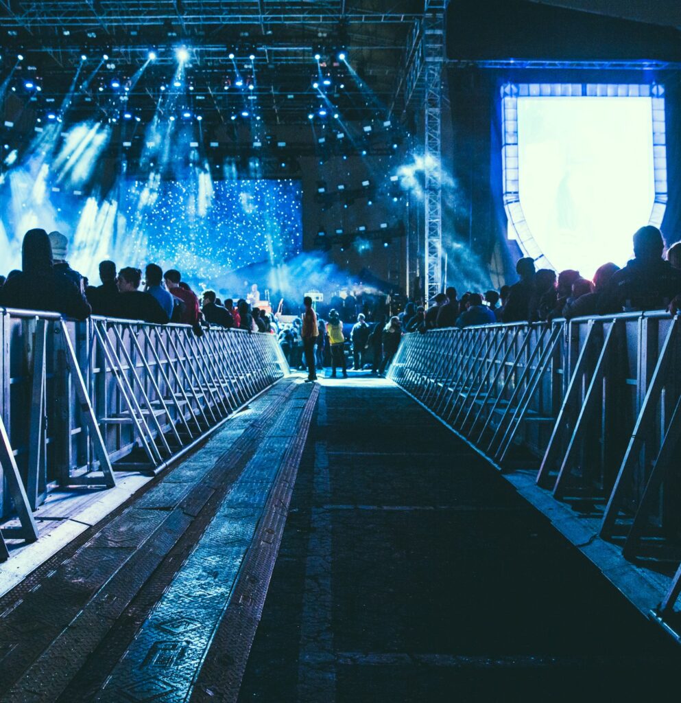 Concert scene with a crowded audience. Blue stage lights illuminate the area, casting a lively and energetic atmosphere. Barriers line the walkway.