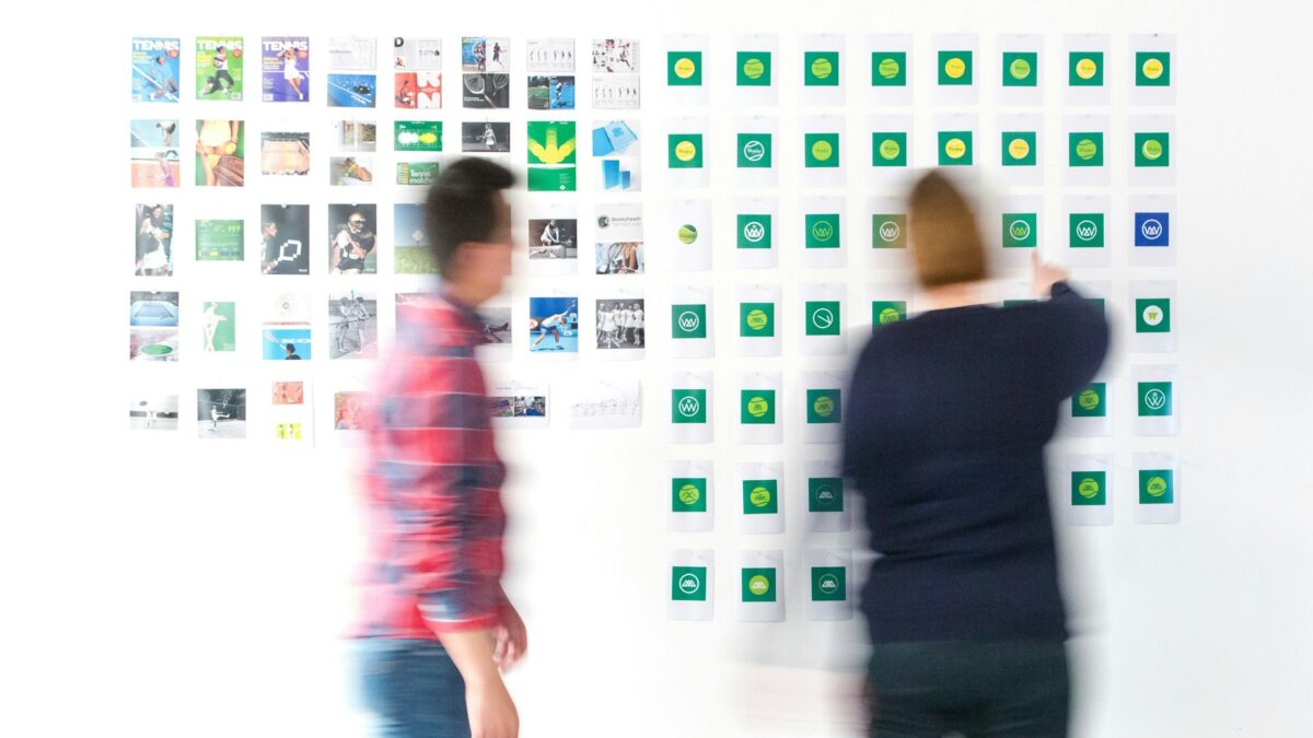 Two blurred figures stand before a wall of colorful photos and symbols, suggesting brainstorming or creative planning