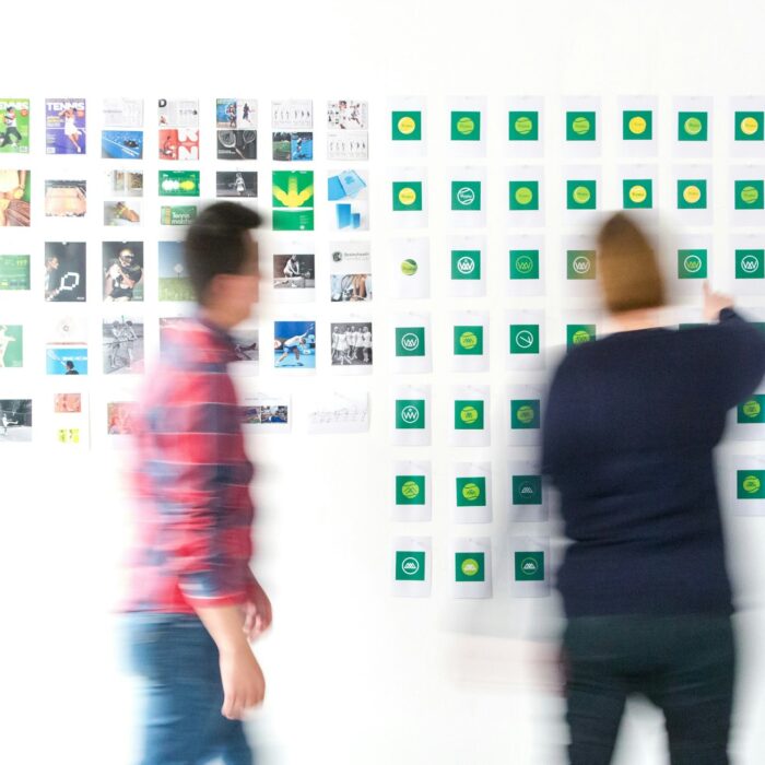 Two blurred figures stand before a wall of colorful photos and symbols, suggesting brainstorming or creative planning