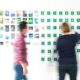 Two blurred figures stand before a wall of colorful photos and symbols, suggesting brainstorming or creative planning