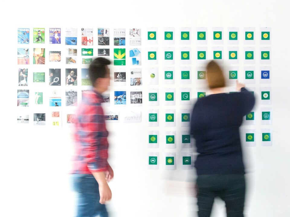 Two blurred figures stand before a wall of colorful photos and symbols, suggesting brainstorming or creative planning