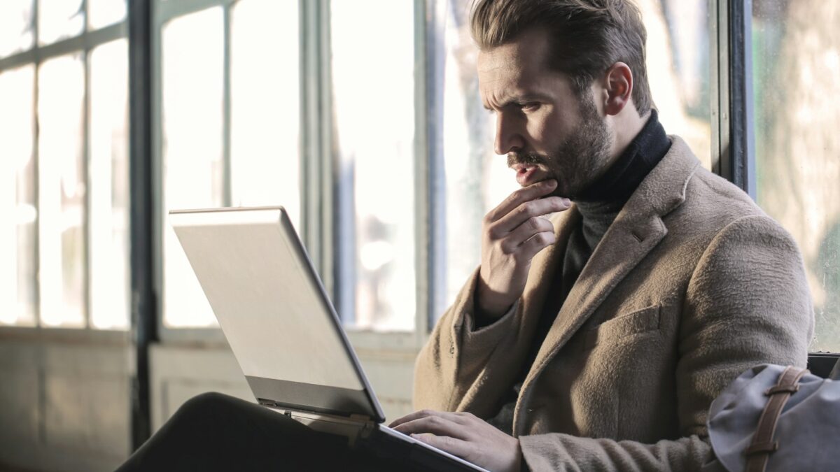 A man in a brown coat sits by the window, focused on a laptop. He touches his chin thoughtfully.