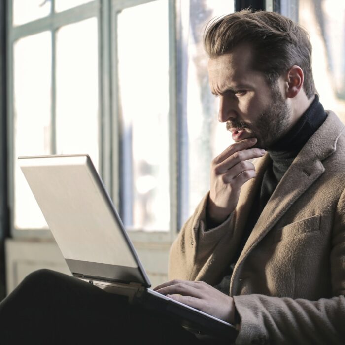 A man in a brown coat sits by the window, focused on a laptop. He touches his chin thoughtfully.