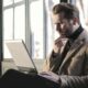 A man in a brown coat sits by the window, focused on a laptop. He touches his chin thoughtfully.