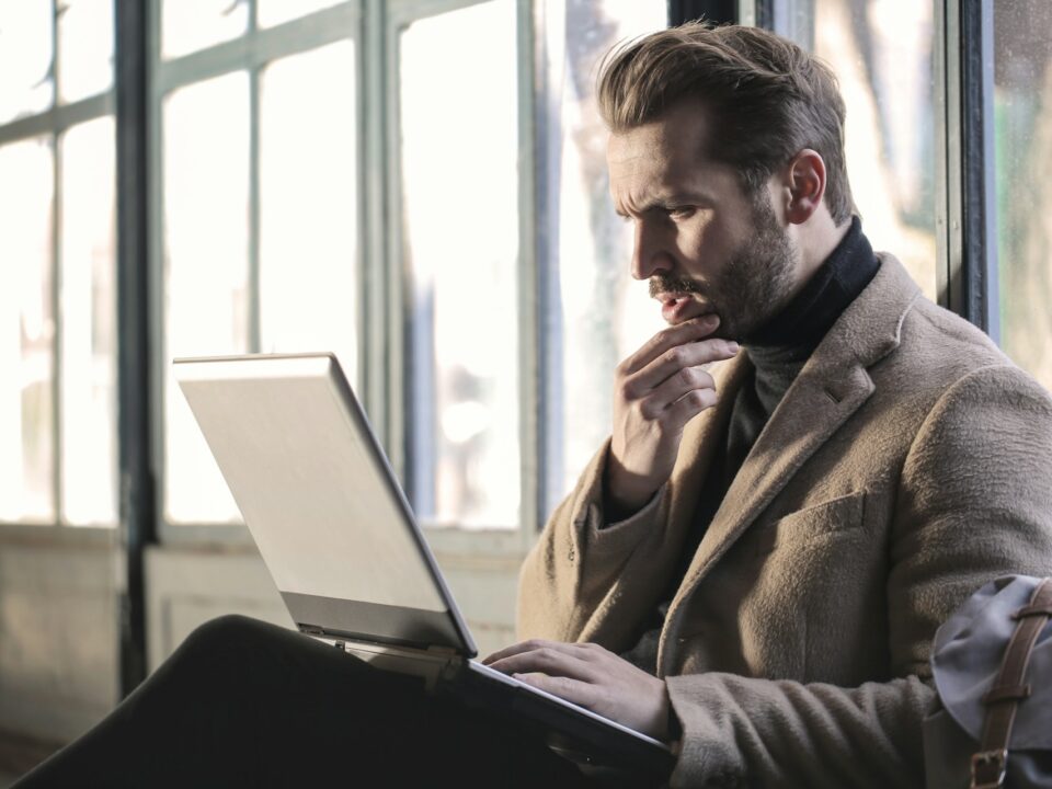 A man in a brown coat sits by the window, focused on a laptop. He touches his chin thoughtfully.