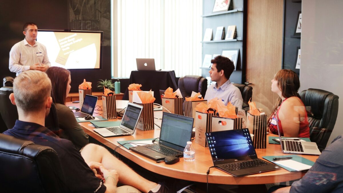 A group of seven people sit around a conference table with laptops, listening to a presenter in a meeting room.