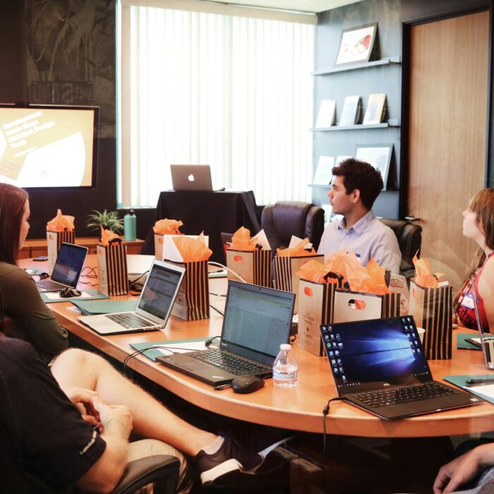 A group of seven people sit around a conference table with laptops, listening to a presenter in a meeting room.