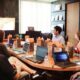 A group of seven people sit around a conference table with laptops, listening to a presenter in a meeting room.