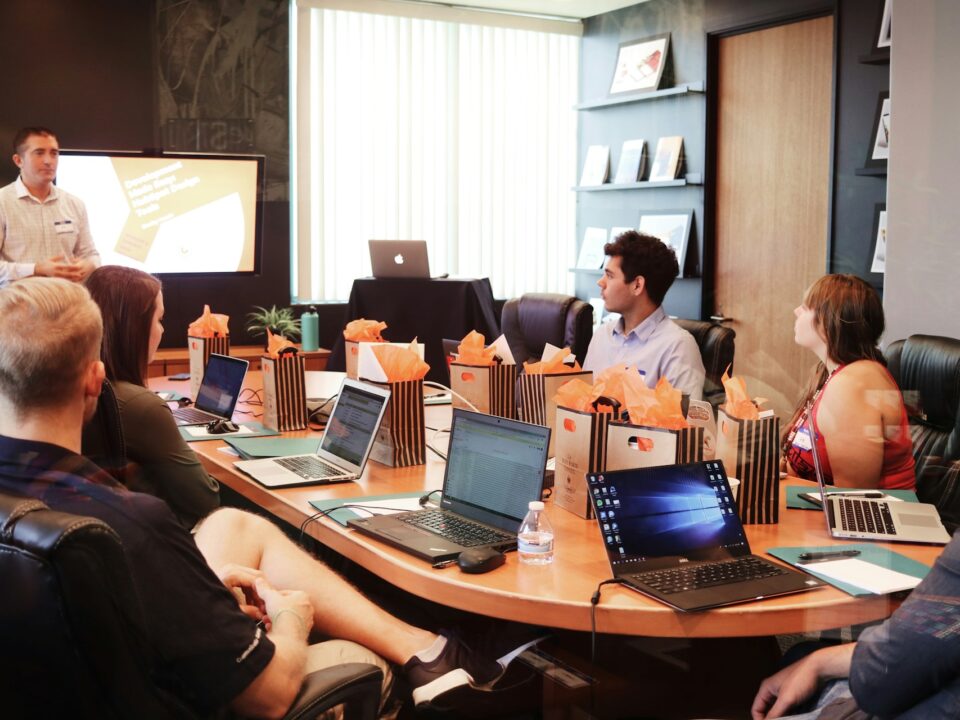 A group of seven people sit around a conference table with laptops, listening to a presenter in a meeting room.