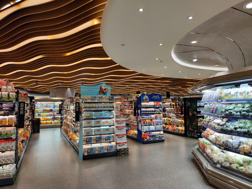 Modern supermarket interior with vibrant lighting and wavy ceiling design. Shelves are stocked with groceries and fresh produce