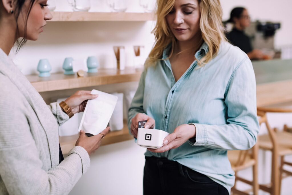 Two women in a cafe, one in a light blue shirt holding a credit card reader, processing a purchase for the other. Shelves with mugs behind.