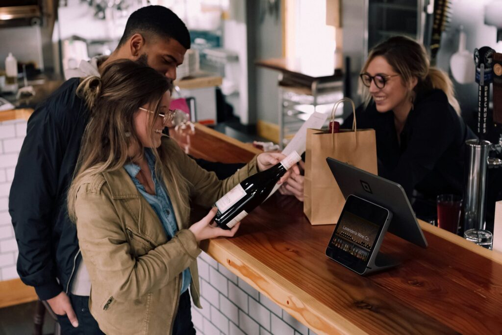 A woman with glasses and a man are at a bar counter, smiling as they hold a bottle of wine, interacting with a cashier who is handing them a receipt.