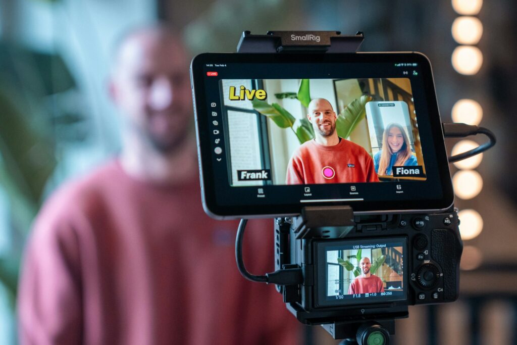 A live streaming setup shows a close-up on a monitor with two people labeled "Frank" and "Fiona" smiling, with blurred bokeh lights in the background.
