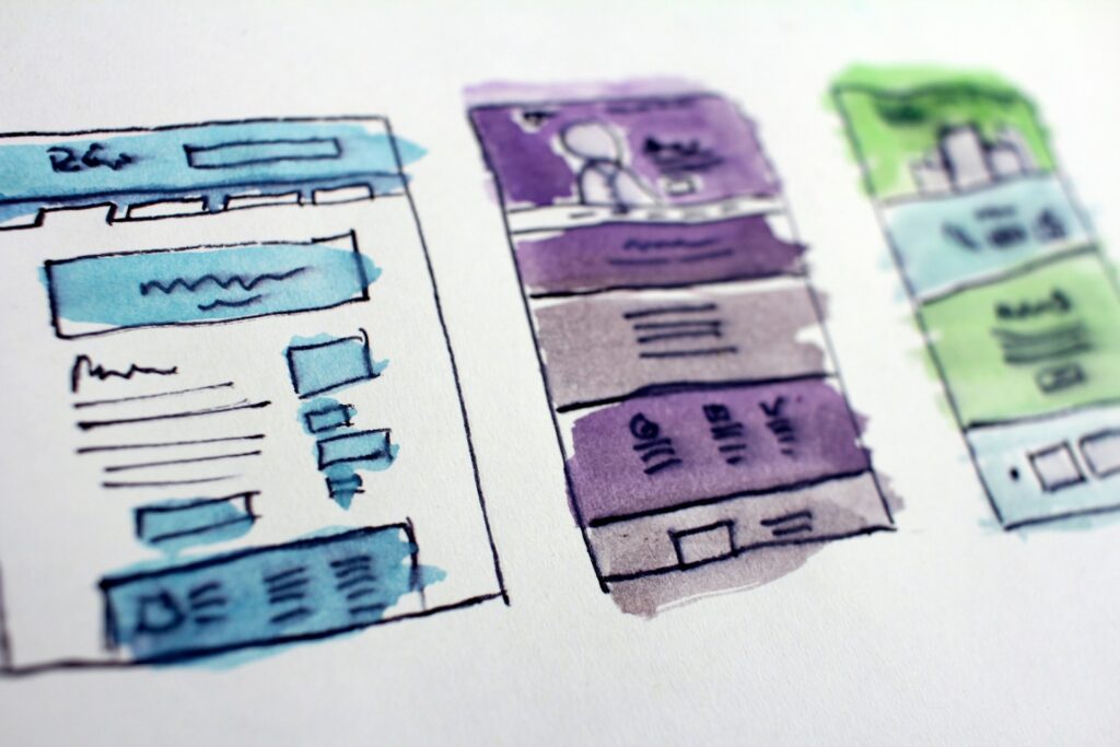 Hand-drawn sketches of three colorful website templates on paper, with blue, purple, and green sections. They suggest creative web design ideas.