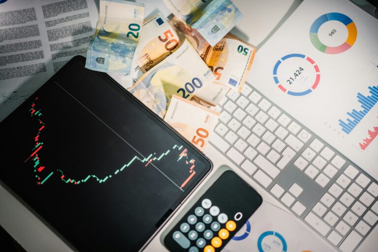 A desk with Euro banknotes, a keyboard, graphs, and charts on paper, alongside a tablet showing a stock market graph and a calculator.