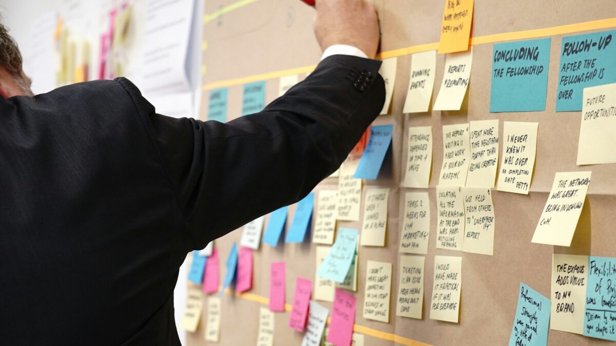 A person in a suit writes on a brown board filled with colorful sticky notes.