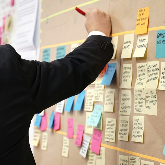 A person in a suit writes on a brown board filled with colorful sticky notes.