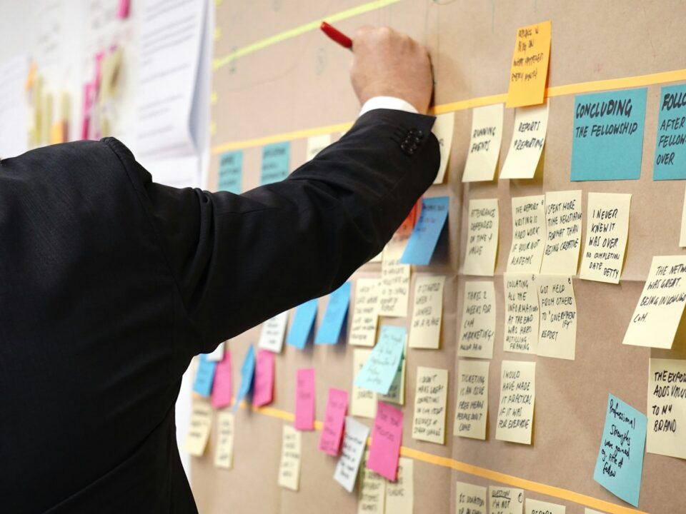 A person in a suit writes on a brown board filled with colorful sticky notes.