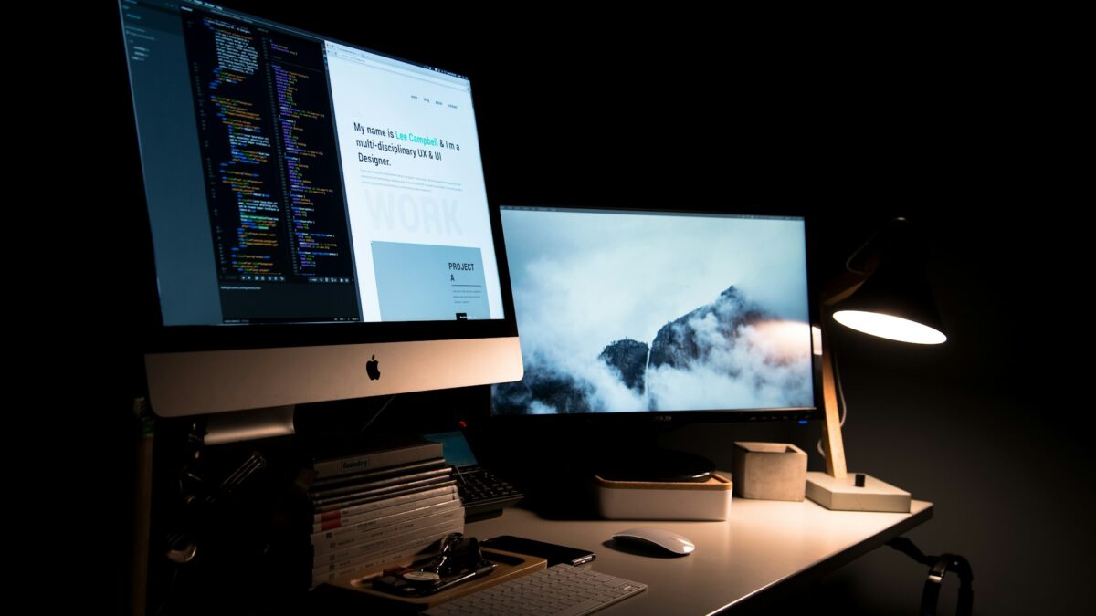 Dimly lit workspace with two monitors on a desk in a dark room. One screen shows code, the other displays a foggy mountain scene.