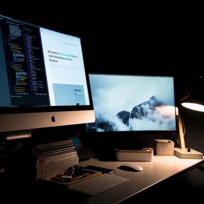 Dimly lit workspace with two monitors on a desk in a dark room. One screen shows code, the other displays a foggy mountain scene.