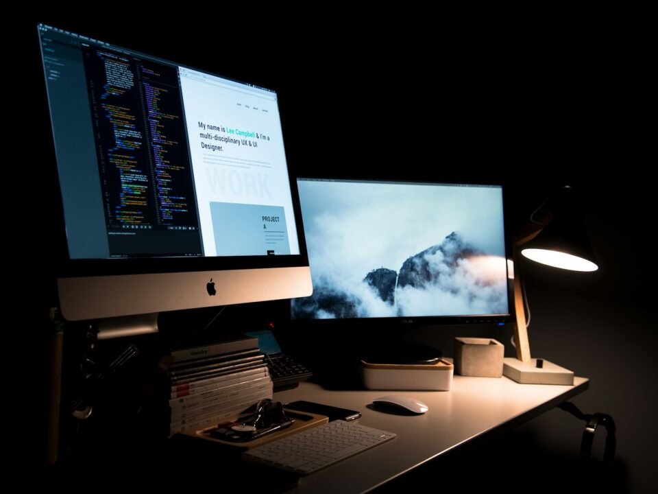 Dimly lit workspace with two monitors on a desk in a dark room. One screen shows code, the other displays a foggy mountain scene.