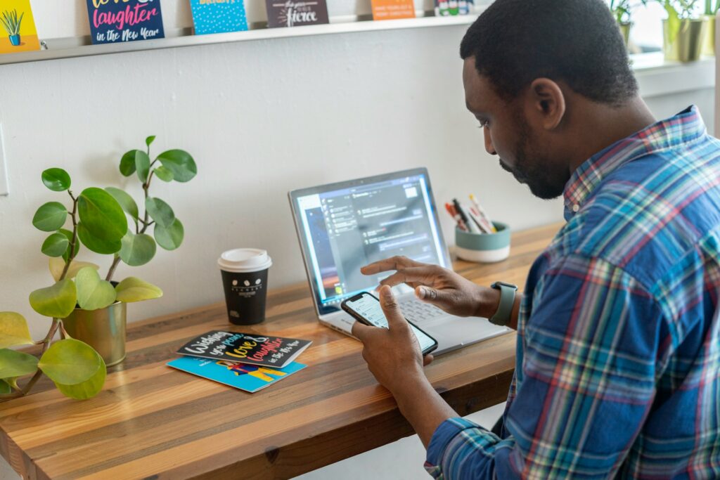 A man in a plaid shirt uses a smartphone while seated at a wooden desk. A laptop, coffee cup, plant, and colorful cards are nearby