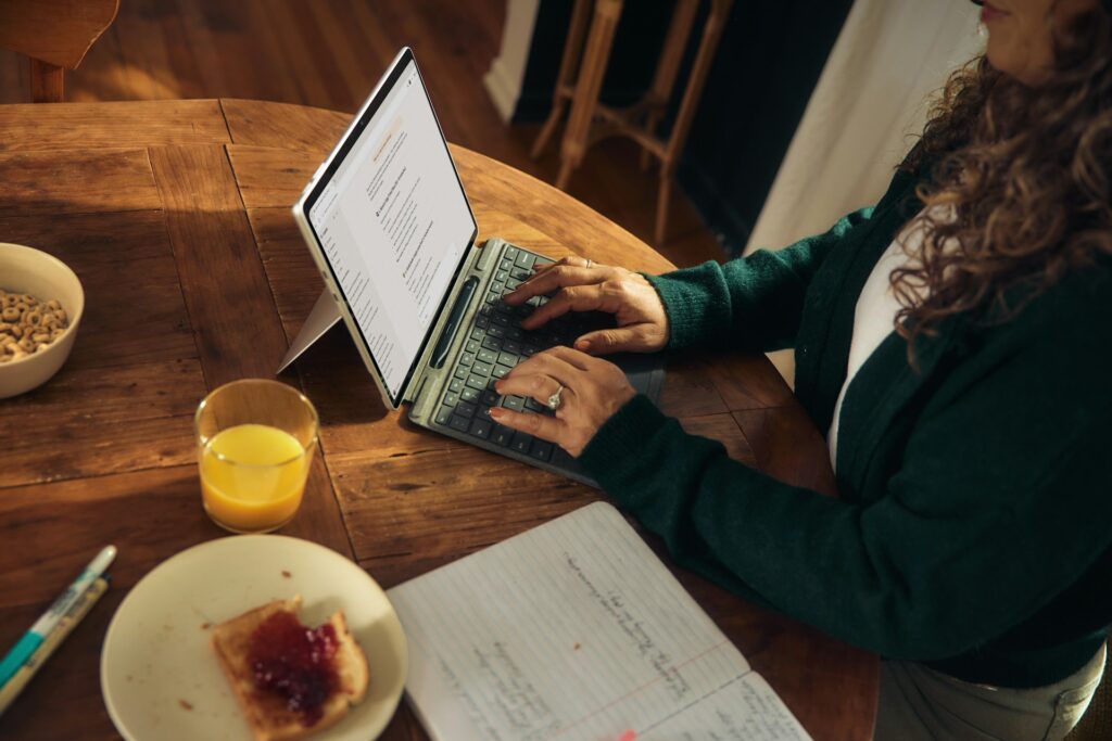 A person types on a laptop at a wooden table, surrounded by breakfast items like toast with jam, cereal, and orange juice. A notebook lies nearby.