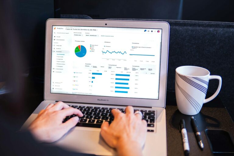 Person using a laptop showing analytics with graphs and charts. A coffee cup is nearby, with pens on the desk.