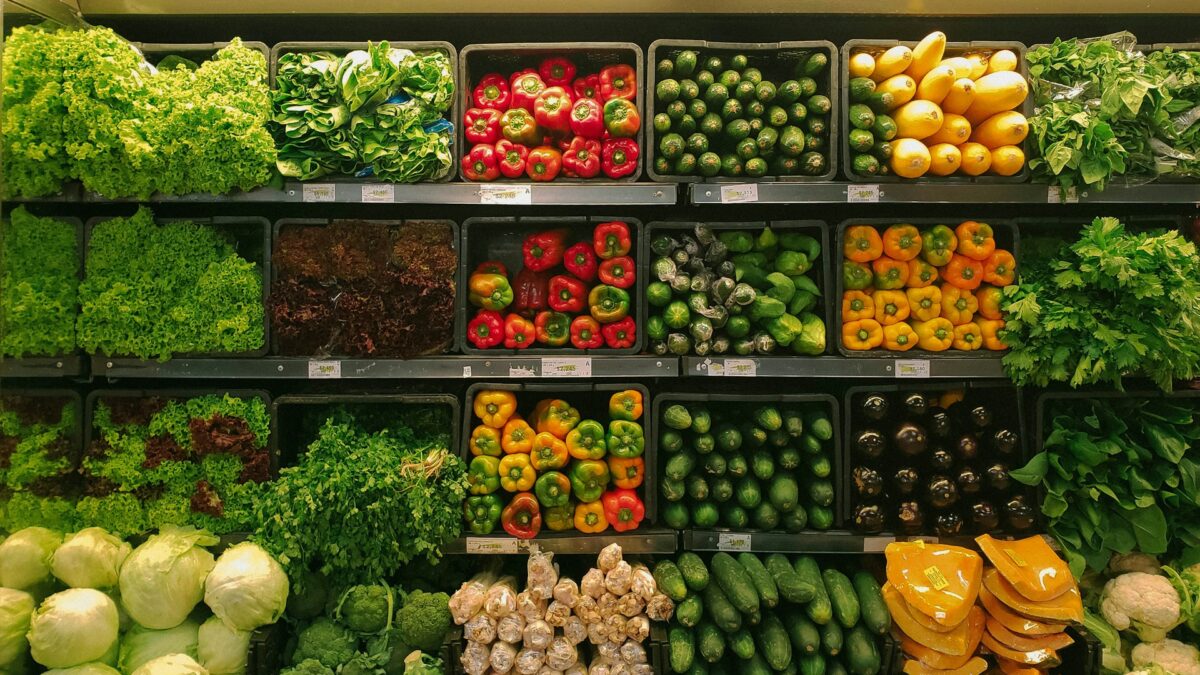 A vibrant supermarket vegetable display features green lettuce, red and yellow bell peppers, cucumbers, squash, and leafy greens neatly arranged on shelves.