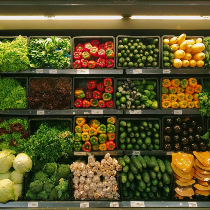 A vibrant supermarket vegetable display features green lettuce, red and yellow bell peppers, cucumbers, squash, and leafy greens neatly arranged on shelves.