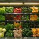 A vibrant supermarket vegetable display features green lettuce, red and yellow bell peppers, cucumbers, squash, and leafy greens neatly arranged on shelves.