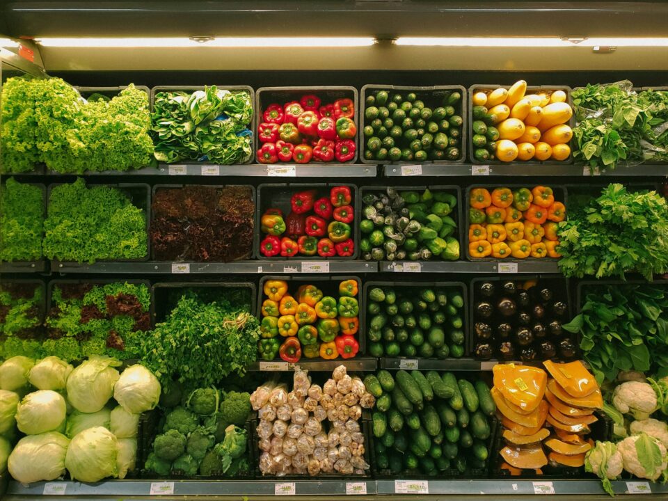 A vibrant supermarket vegetable display features green lettuce, red and yellow bell peppers, cucumbers, squash, and leafy greens neatly arranged on shelves.