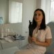 A woman in a white uniform sits in a clinic room, putting on a black glove. Skincare products are on a nearby table.