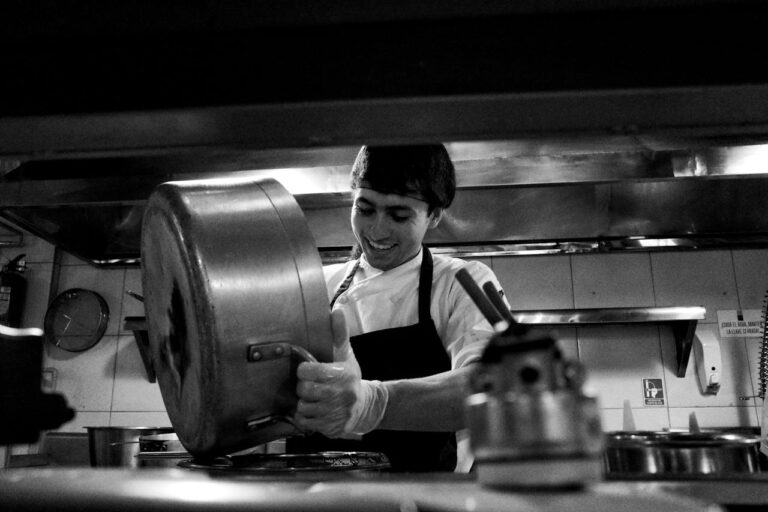 A chef smiles while pouring food from a large pot in a commercial kitchen.