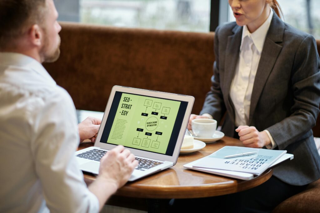 Two professionals discuss an SEO strategy at a cafe. A laptop displays a flowchart. They are dressed formally, each holding a cup of coffee.