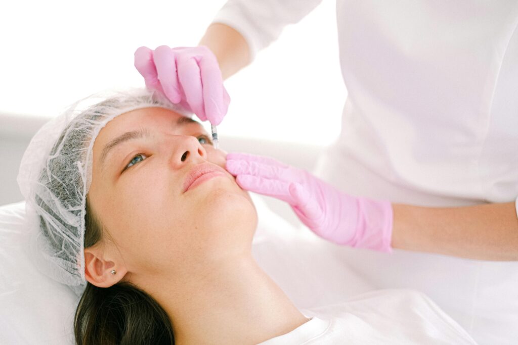 A woman receives a beauty treatment, lying down with her hair covered. A professional in pink gloves gently doing the procedure.