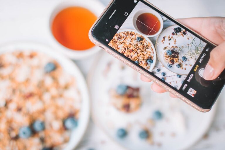 A hand holds a smartphone capturing a top-view shot of a breakfast setup, featuring granola with blueberries and a cup of tea.