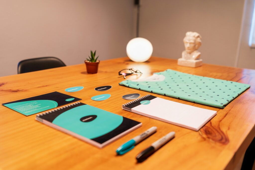A tidy office desk displays a teal-themed stationery set with notebooks, pens, and stickers. Nearby, a plant, glasses, and a glowing lamp add ambiance.
