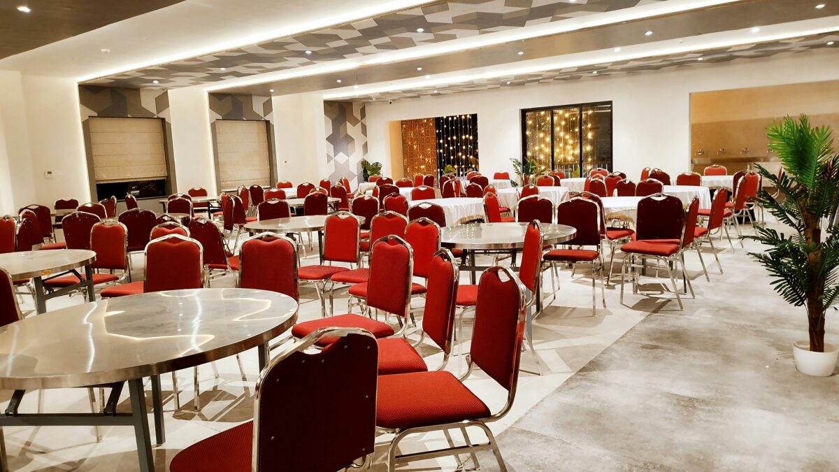 Spacious, elegantly lit banquet hall with round tables, red chairs, and potted plants.