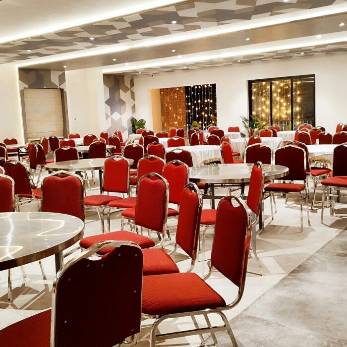 Spacious, elegantly lit banquet hall with round tables, red chairs, and potted plants.
