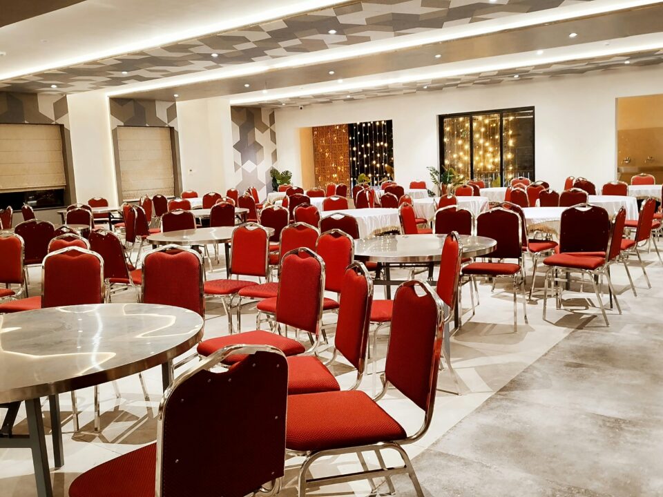 Spacious, elegantly lit banquet hall with round tables, red chairs, and potted plants.