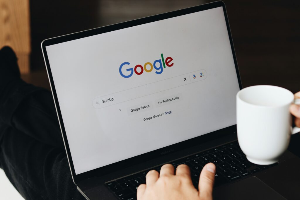 Person using a laptop displaying Google's search page, typing "SumUp" in the search bar while holding a white coffee cup