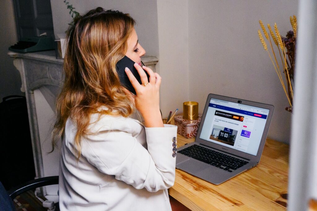 A woman with wavy hair in a white blazer talks on a phone while working on her laptop at a wooden desk.