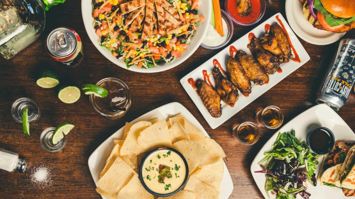 A table with colorful dishes, including a salad, chicken wings, a burger, nachos with dip, and drinks.